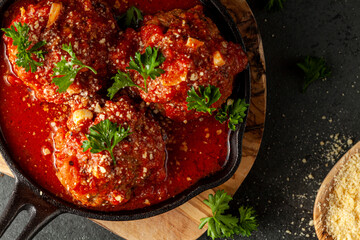 Close up view of three traditional large Italian meatballs in sizzling tomato sauce in cast iron pan. The pan sits on wooden trivet and parsley leaves and grated Parmesan Cheese is used for flavor