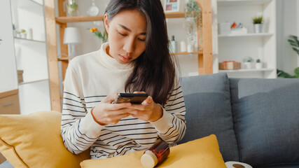 Sick young Asian woman hold medicine sit on couch take photo sent to doctor at home. Girl take medicine after doctor order, quarantine at house, Coronavirus social distancing quarantine concept.