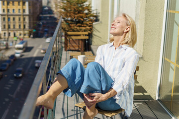 Enjoying morning. Beautiful dreamy mature 60s woman in casual wear relaxing on balcony at home, starting new day with coffee. Happy elderly woman sitting on terrace and looking at sunny street