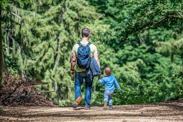 father holding hand of little son with backpack hiking in forest