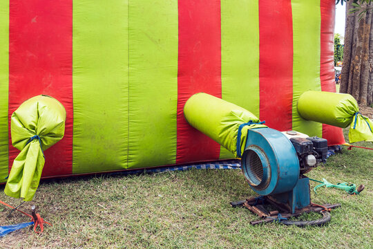 The old portable air blower pump fan install with green and red inflatable bouncer ball house castle or toy. Outdoor blue electric blower in the playground. Motor Air Blower on lawn in kid play land.