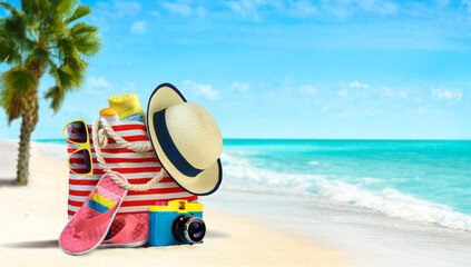 Summer beach bag and accessories - straw hat, flip flops and sunglasses on sandy beach and azure...