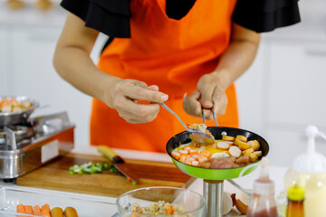 Mom wearing an orange apron in home kitchen learning to cook family food of hotdogs, crab sticks, hams for kid by using spoon to pour healthy salad into flying pan