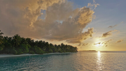 Sunrise in the Maldives. The sky and clouds are highlighted in shades of pink and gold. Sunny path on the ocean surface. On the shore there are thickets of palm trees. Silence and serenity.