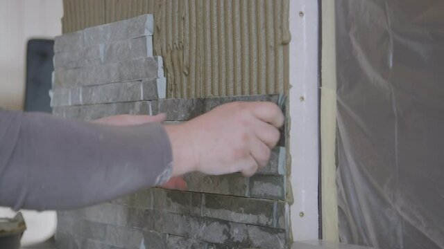 SLOW MOTION, CLOSE UP: Builder presses a tile firmly against the wall covered in ridged mortar. Unrecognizable tiler fits a natural stone tile on the living room wall. Decorating the house interior.