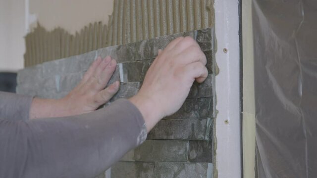 SLOW MOTION, CLOSE UP: Unrecognizable tiler fits a natural stone tile on the living room wall. Builder presses a tile firmly against the wall covered in ridged mortar. Decorating the house interior.