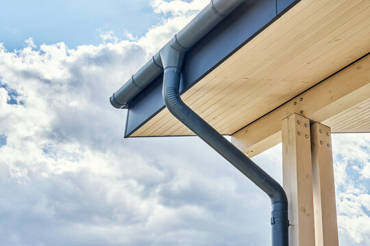 Contemporary grey metal rainwater downpipe installed on roof of new building with wooden terrace on cloudy day close view