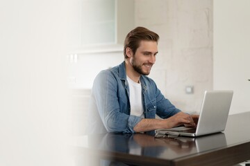 Young casual man working on laptop online, sitting at table in kitchen, looking at computer screen