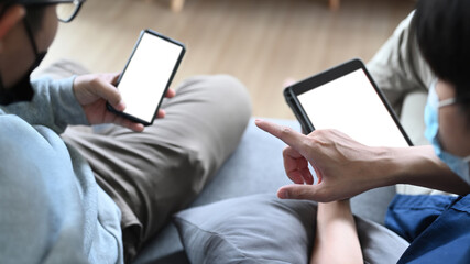 Close up view of two casual man wearing protective mask using digital tablet while sitting on sofa.