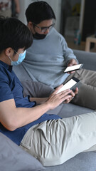 Two man wearing protective mask using smart phone and sitting together on sofa.