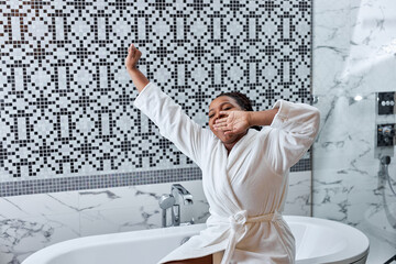 Woman In Bathroom. African lady in bathrobe sits on bathtub in the morning. Woman is going to take bath after waking up, yawning. In Bright cozy bathroom - obrazy, fototapety, plakaty