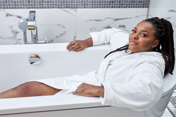 Young african model in bathrobe sitting inside of white bath, preparing relaxing bath. Beauty, healthy lifestyle concept. dark skinned lady have rest alone at home in the morning - obrazy, fototapety, plakaty