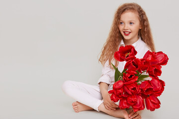 Happy little blonde girl looking away with toothy smile and positive facial expression, sitting on floor, holding bouquet of red tulips, isolated over gray background.