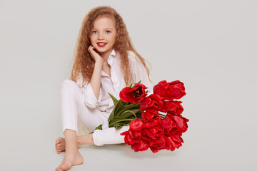 Cute confident little schoolgirl sitting on floor, looking smiling at camera, keeping hand under chin, holding bouquet of red tulips, isolated over gray background.