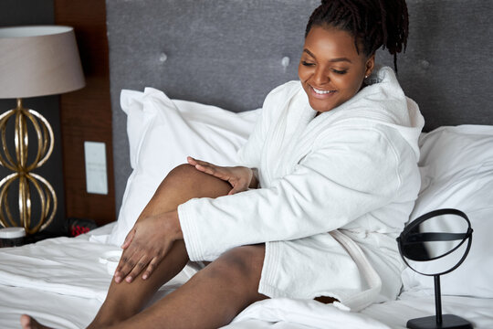 Black woman in bathrobe touching soft legs after epilation and depilation procedure, spending morning alone, self-care concept. at home