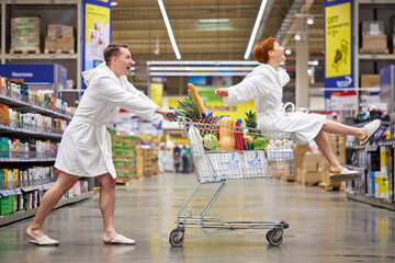 Man and woman in white bathrobes having fun riding shopping cart and sitting on it, man holding...