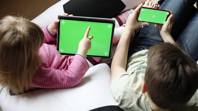 Kid boy and little girl gaming or using social media on mobile phone and tablet with chroma key background. Two children holding smartphone and laptop with green screen for commerce, new app