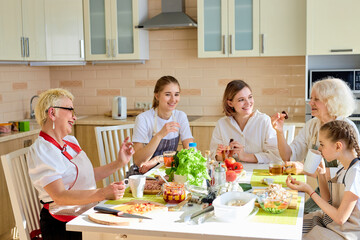 Optimistic family women and children drinking tea after cokking, side view on beautiful females having rest, sitting at table enjoying time together, have talk, laughing. at home, in bright kitchen