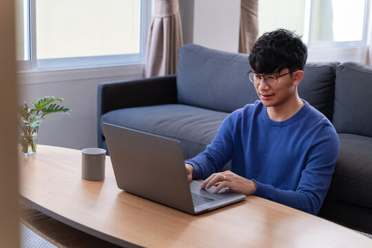 Young Attractive Asian Man Relaxing Comfortably Working On Laptop In Bright Living Room At His Home.