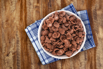 Top view of bowl with sweet chocolate cereal flakes for breakfast on white wooden background