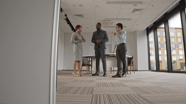 Tracking right of two multiethnic businessmen and mixed-race businesswoman standing together in modern office and discussing future company strategy