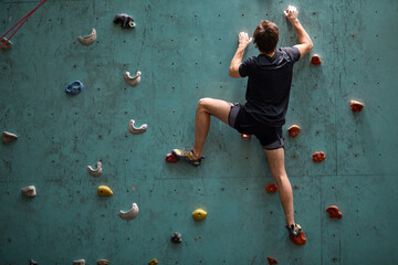 active young caucasian man on rock wall in sport center, rock climbing. Sportive male guy in black sportswear engaged in sport, fitness, lead healthy lifestyle. Rear view - obrazy, fototapety, plakaty