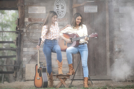Two Asian women have long hair in country-style clothes standing on a farm with a guitar.
