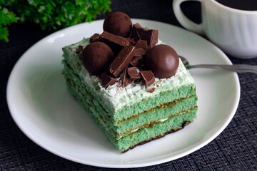 Delicious rectangular green cake on a white plate decorated with pieces of chocolate and brown candies. Sweet cake on a dark background.