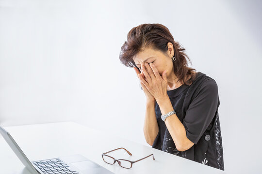 パソコンを前に眼の疲れに顔を覆うシニア女性。疲れ,疲労,過労イメージ