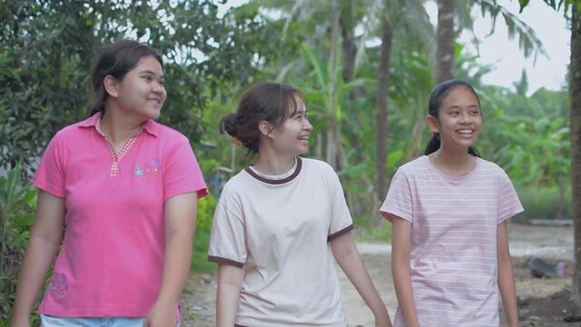 Cheerful three Thai teen girls in casual dress talking and walking leisurely together on the footpath in the rural village. Happy healthy female teenager lifestyle. Asian siblings.