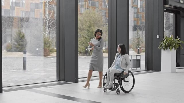 Panning of beautiful handicapped businesswoman in wheelchair and mixed-race businesswoman going along hall of office center and having chat