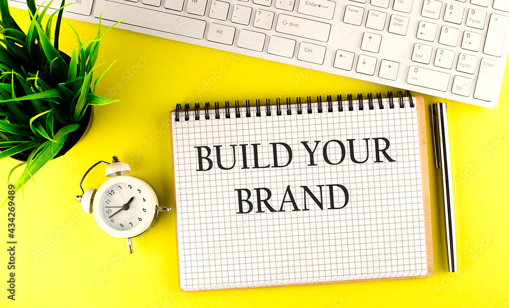 Poster build your brand text on notebook with keyboard , pen and alarm clock on the yellow background