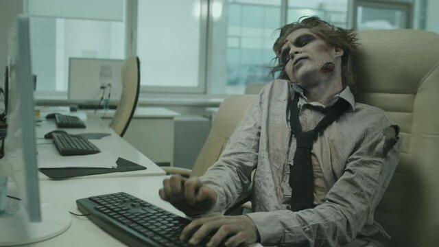 Medium shot of zombie office worker sitting at desk and smashing keyboard with his hands while trying to type