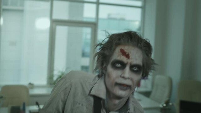 Handheld Tracking Shot Of Zombie Man With SFX Makeup And Fake Wounds Wearing Torn Dirty Shirt And Necktie Walking Towards Camera In Empty Office