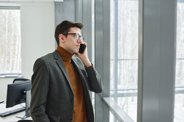 Young businessman in eyeglasses looking through the window and talking on mobile phone at office