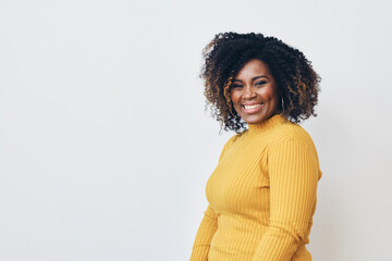 Studio portrait of cheerful mid adult black woman smiling at camera. Curly hair, yellow sweater, white background. 