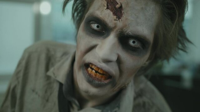 Handheld close up of zombie man with white contact lenses and SFX makeup with fake wounds looking at camera and grunting