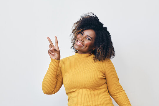 Studio portrait of cheerful mid aged black woman with curly hairstyle making peace sign with right hand.