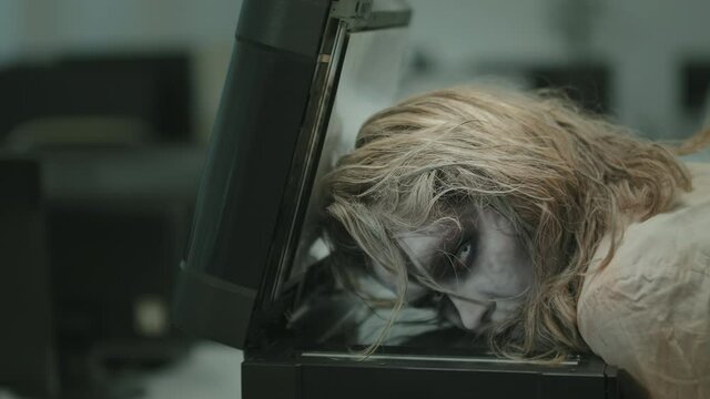 PAN shot of young zombie woman with white contact lenses and SFX makeup lying on copier in office