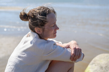 Woman in linen clothes on stone sitting near sea, looking to water. Wellbeing, mental health. Hugging herself