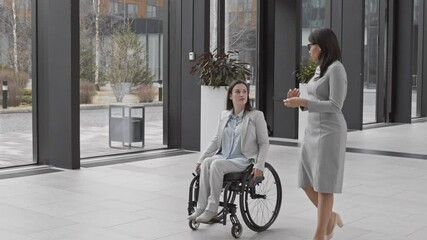Pan shot of Caucasian attractive handicapped businesswoman moving in wheelchair along office center hall while talking to beautiful mixed-race businesswoman going nearby - Powered by Adobe