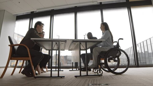 Wide shot of two multiethnic businesswomen and two businessmen sitting at desk in office with panoramic windows and brainstorming during discussion of future company strategy