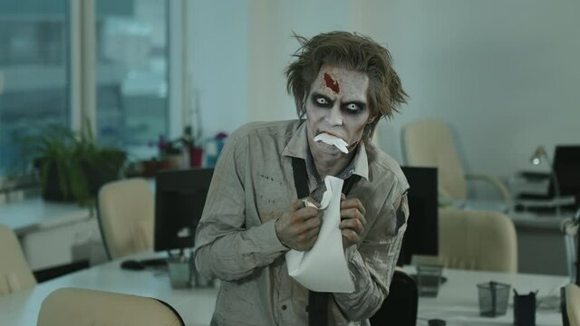 Portrait shot of zombie man with contact lenses, SFX makeup and fake wound on forehead looking at camera and eating document in office