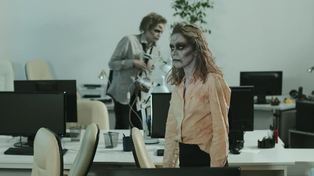 PAN slowmo of zombie businesswoman and businessman wearing SFX makeup, contact lenses and dirty clothes walking between desks in empty office