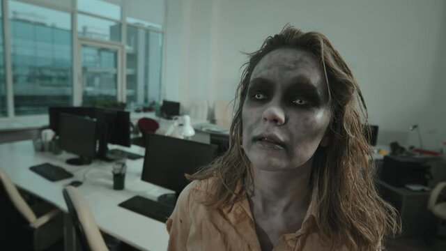 Close up of mindless female zombie with contact lenses and scary SFX makeup looking at camera in office