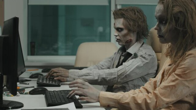 Side view shot of mindless zombie office workers in dirty and torn clothes sitting at desks and typing on keyboards