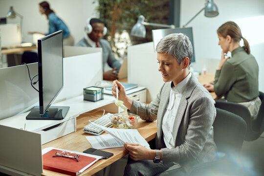 Smiling female entrepreneur reading business reports while eating salad on lunch break in the office. - Powered by Adobe