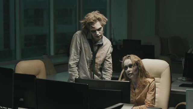 PAN slowmo shot of twitching zombie businessman in torn shirt and necktie standing by desk of female zombie colleague working on computer in dark office