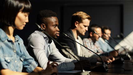 Side view of group of multiethnic business people sitting at desk in a row taking part in conference, microphones in front of them. Focus on young Black man speaking