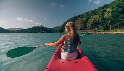 Long haired blonde woman with sunglasses rows bright pink canoe along sea bay water to beach with growing palms.